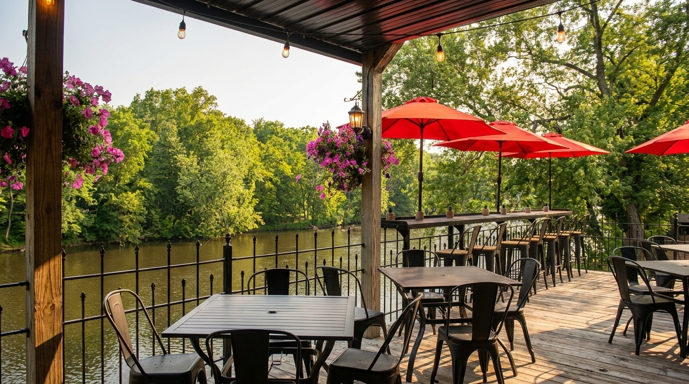 Outdoor restaurant patio overlooking a river, with black tables, chairs, and red umbrellas.
