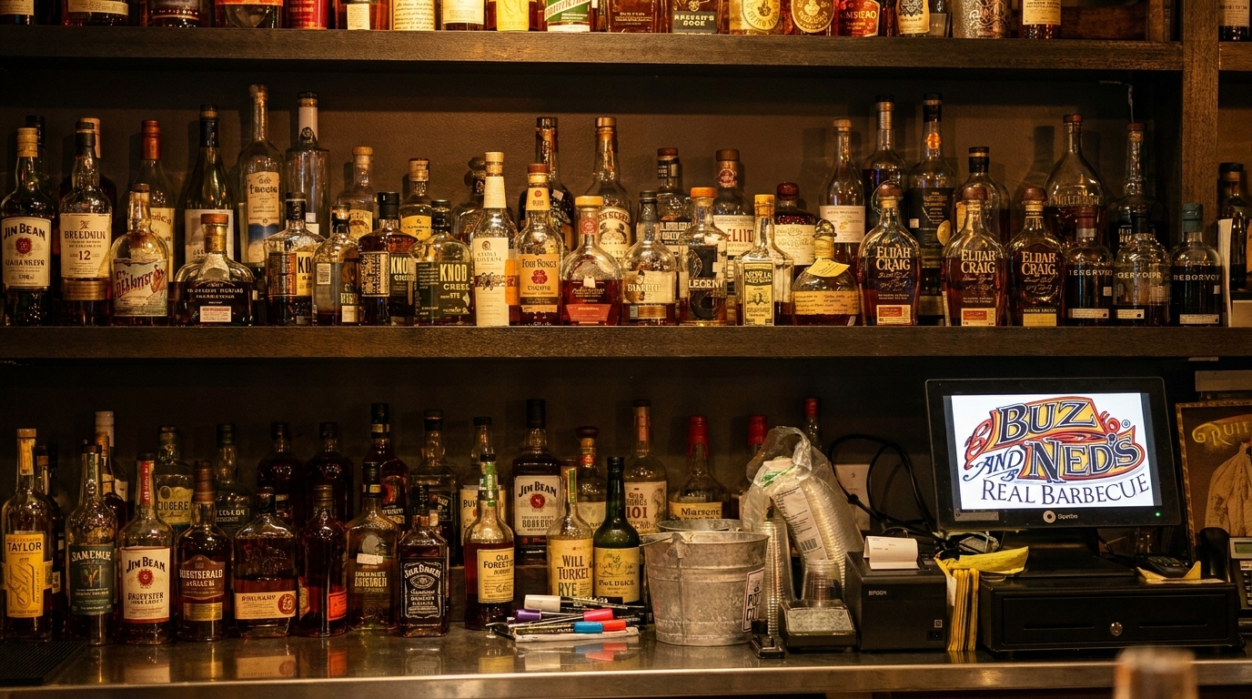 A well-stocked bar shelf with numerous bottles of liquor and a monitor displaying "Buz and Ned's Real Barbecue".
