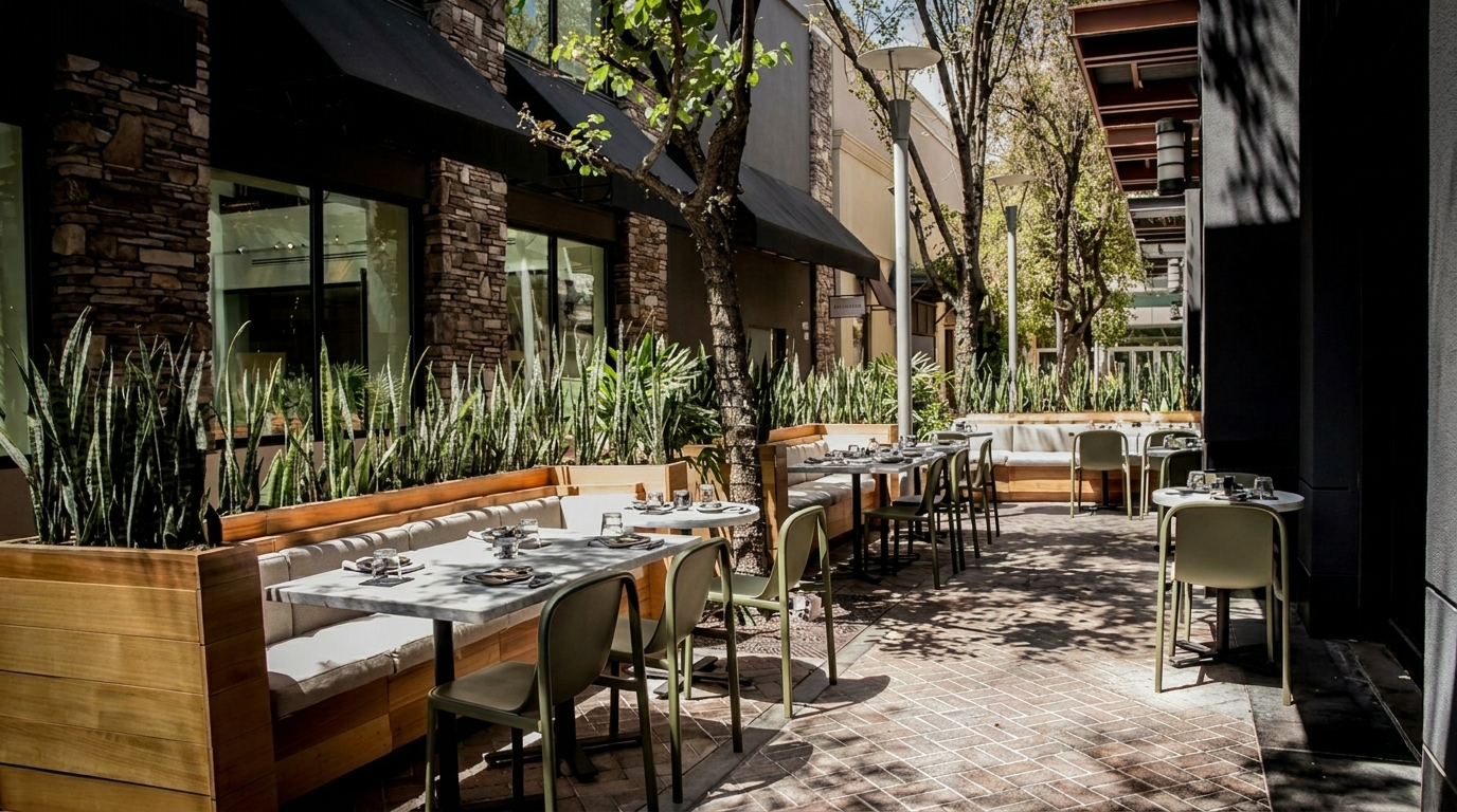 Outdoor patio dining area with tables, chairs, and benches, surrounded by lush green plants and trees.
