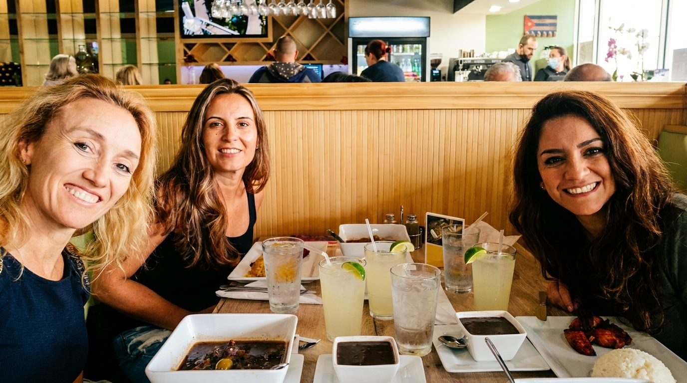 Three smiling women at a restaurant table with Cuban food and drinks, other diners in background.