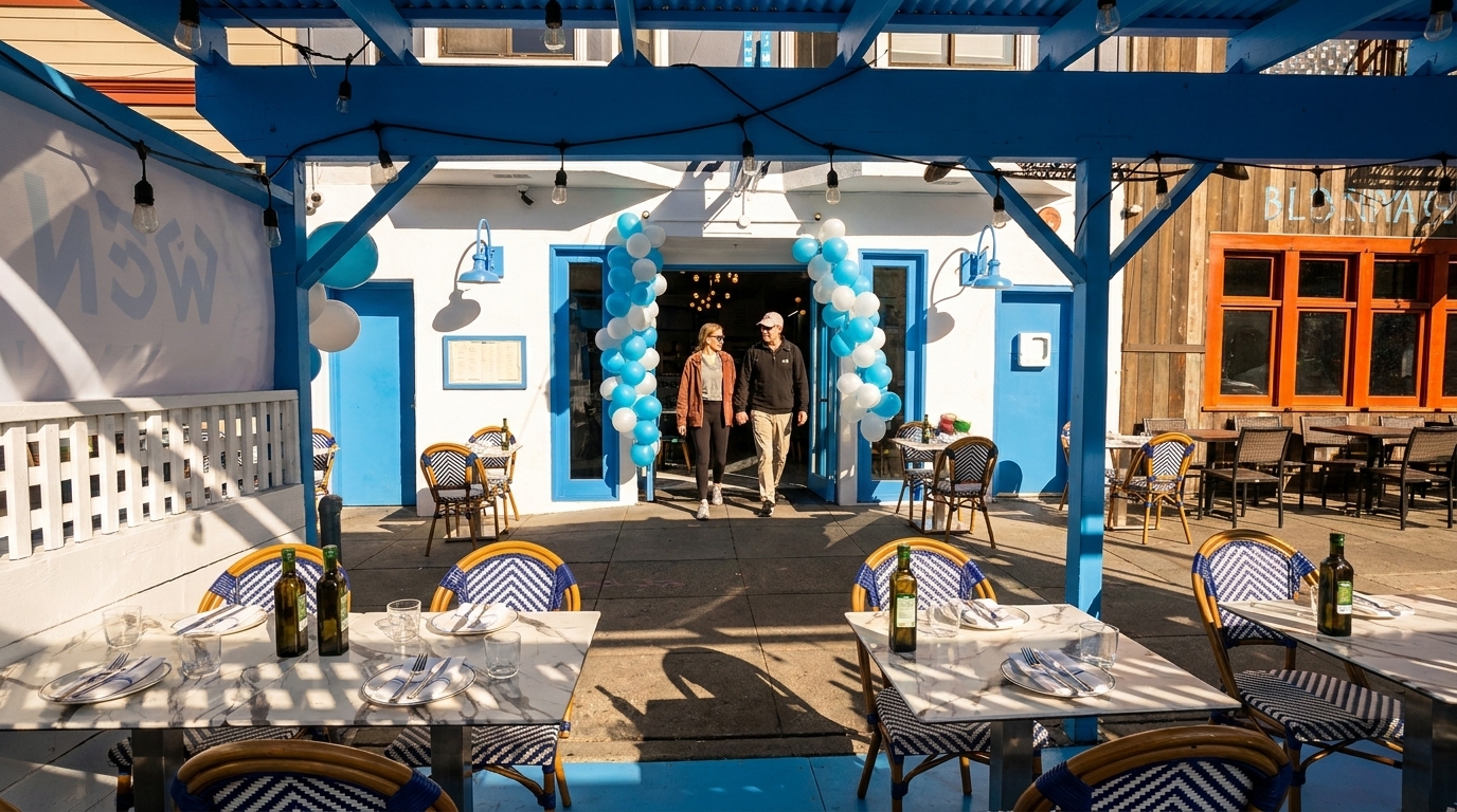 Two people walk out of a restaurant entrance decorated with blue and white balloons, past outdoor dining tables.